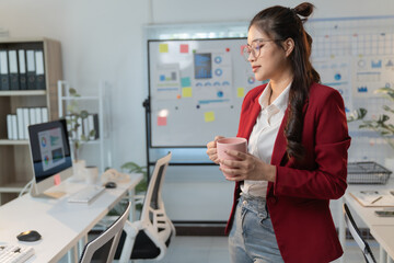 Young businesswoman drinking from a pink mug and thinking about work while standing in a modern office with a whiteboard with graphs and charts in the background