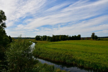 A view of a shallow yet vast pond or lake flowing next to vast fields covered with herbs, reeds, and various crops located next to some tiny forests and moors spotted on a cloudy summer day in Poland
