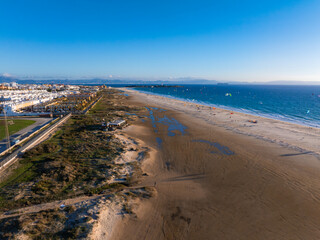 Tarifa, Spain, features a sandy beach with kite surfers and colorful kites. White buildings line the coast, with mountains visible on the horizon.