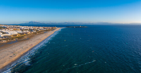 Tarifa, Spain, features sandy beaches, white buildings, and kite surfers. A lighthouse stands on a peninsula, with Rif Mountains visible across the strait.