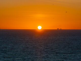 Fototapeta premium The sun sets on the horizon near Tarifa, Spain, casting an orange glow on the ocean. A large ship is visible in the distance, with birds flying above.