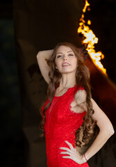 Fototapeta premium Fire and Grace: A young beauty in a red dress posing against a backdrop of flames