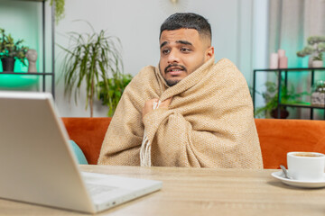 Sick ill Indian man freelancer working on laptop wrapped in plaid sit shivering from cold drinking...