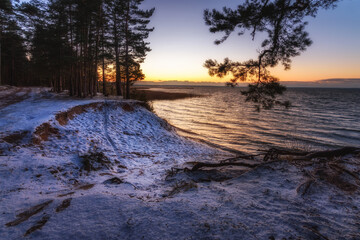 Evening sunset over a large lake with a wave and a high snow-covered coast with a cliff, basin and pine trees. Beautiful winter landscape. side view from the shore