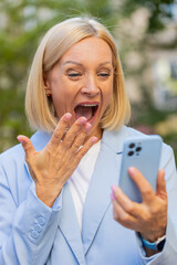 Happy shocked excited Caucasian mature businesswoman in formal suit mouth opened celebrate victory, achievement holding mobile phone good news win message rejoice business success outside city street.