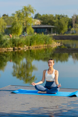 Young woman sitting on mat in lotus position, relaxing near pond in park on summer day. Sporty...
