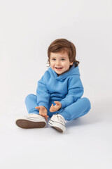 Naklejka premium A Caucasian curly-haired toddler boy of 3 years old is sitting on the floor, smiling and looking attentively with interest in a blue tracksuit on a white background. Vertical photo. Copy Space