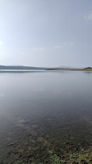 Serene lake with still waters, grassy shorelines, and hazy distant hills under a clear blue sky