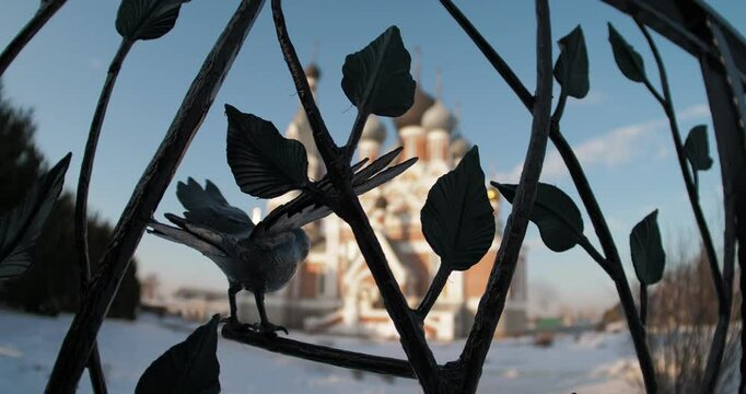 Religion, architecture. The figure of a bird in a wrought-iron fence against the background of a Christian temple is out of focus on a cold day. Hand held, distortion . Siberia.