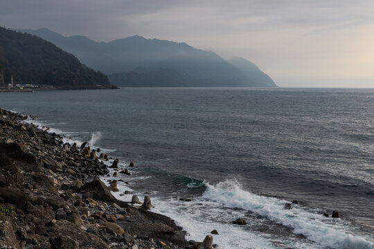 Waves crash against a rocky shoreline with mist-shrouded mountains in the background under an overcast sky.