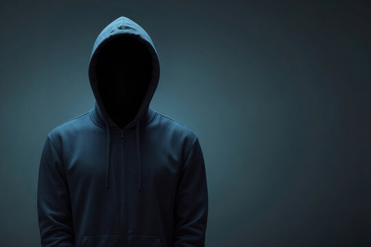 Hacker man is wearing a hoodie and is standing in front of a dark blue background with copy space. Concept of mystery and anonymity, as the man's face is hidden behind the hood