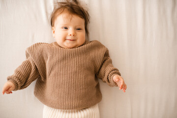 Fototapeta premium Smiling baby girl 5-6 months old lying in bed on white blanket wearing knitted sweater close up. Childhood.