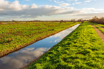 Dutch polder landscape with a straight ditch. It is a windless day in the autumn season and the clouds are reflected in the mirror-smooth water surface.