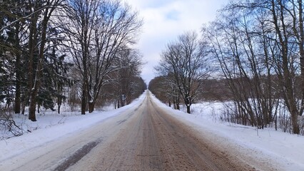 Obraz premium In winter there is snow on the ground, road and trees. The road has not yet been cleared by the road services. There are fields and forest nearby. There are tire tracks on the road. Frosty weather