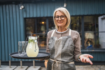 Smile woman waitress serving lemonade outdoors in front blue wall