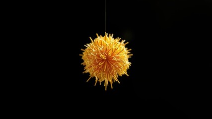 A single golden pom-pom hangs against a stark black background, its delicate strands creating a delicate web of light and shadow.