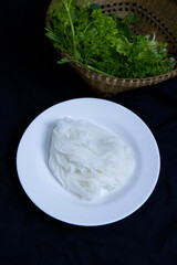 Thai rice flour noodles on white plate with blurred vegetable in rattan basket. Vertical food image of Thai rice noodles for eating, serving, black table setting, traditional food, and Asia concepts.