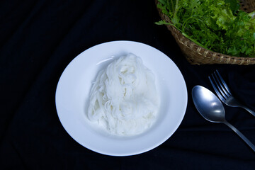 Thai rice flour noodles on white plate, spoon and vegetable basket on black table. Traditional Thai-style table setting of rice flour noodles for eatery, serve, Thai cuisine, and healthy food concept.