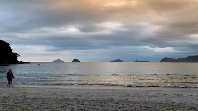 Time-lapse de final de tarde com muitas nuvens e algumas pessoas e cachorros andando na praia. Ao fundo, quatro ilhas: Ilha Mont&atilde;o de Trigo, Ilha dos Gatos, Ilha das Couves e Ilha as Ilhas