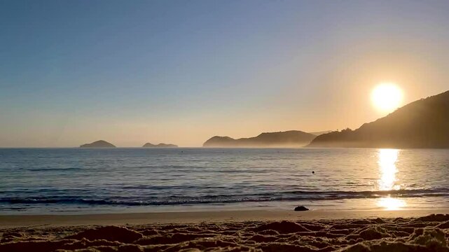 Time-lapse do sol se pondo atr&aacute;s da montanha e seus raios refletindo no mar. V&aacute;rias pessoas, bicicletas, cachorros passando, aparecendo apenas suas silhuetas. Ilha das Couve e As Ilhas ao fundo.