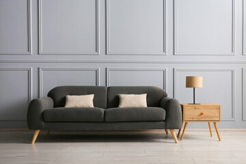 Stylish sofa with cushions and lamp on side table near grey wall