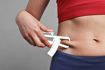 Woman measuring body fat with caliper on gray background, closeup