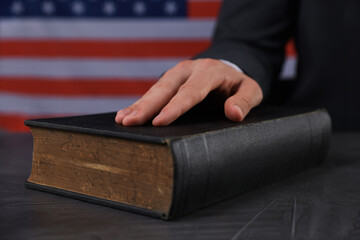 Man taking oath with his hand on Bible at black table against flag of USA, closeup
