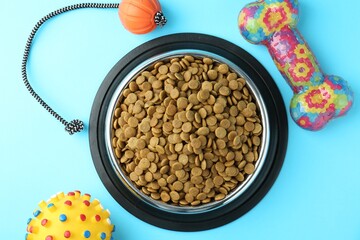Dry pet food in feeding bowl and toys on light blue background, flat lay