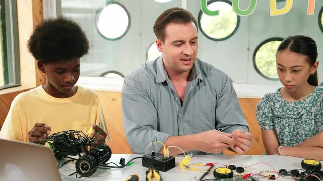 Smart mentor teaching about electronic equipment while fixing car model. Caucasian teacher pointing while explain robotic construction at table with laptop, car model scatter around. Edification