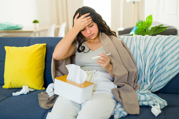 Teenager with cold or flu sitting on sofa at home, wrapped in blanket, checking temperature with thermometer and touching forehead