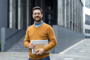 Portrait of a young man standing on the street near an office building, holding a laptop and...