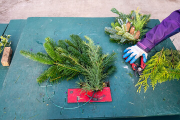 Green wood table with built in red wreath making machine, woman cutting fresh evergreen branch with clippers, celebrate the holidays with handmade decorations
