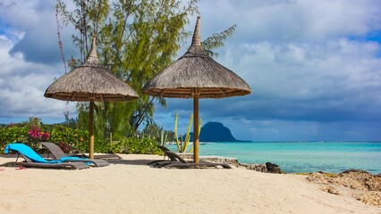 Romantic tropical sandy beach with lounge chairs and straw umbrellas on thunderclouds and blurred Le Morne Brabant peninsula on background. Winter on Mauritius island, Africa