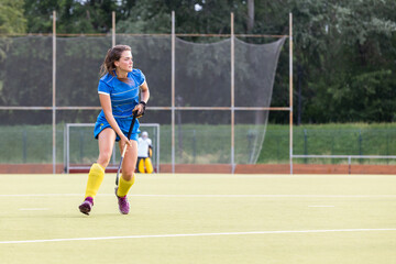 Female athlete in blue uniform running along the pitch during field hockey game