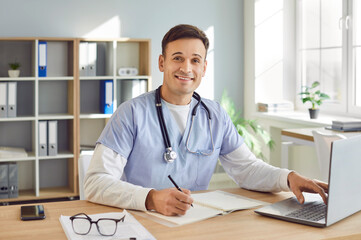 Portrait of cheerful young friendly male doctor in blue medical uniform sitting and working on...
