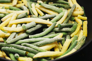 Fried yellow and green string beans with frost on a black pan. Sautéing frozen vegetables for a healthy meal. Close-up of beans with artistic dark contrast. Vegan and vegetarian cooking.