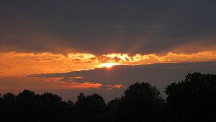 Grey and orange cloudy sky with clouds sun rays during sunset over dark horizon with trees. Topics: air space, weather, meteorology, solar energy, sunlight, natural environment, evening