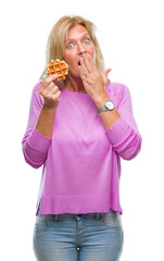 Middle age blonde woman eating sweet waffle over isolated background cover mouth with hand shocked with shame for mistake, expression of fear, scared in silence, secret concept