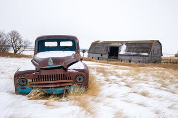 An old, rusted truck is parked in the snow next to a barn © Scott Prokop