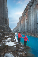 A hiker couple in vibrant red outdoor gear exploring the basalt column formations and turquoise...