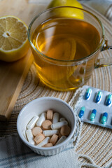 A blister pack of tablets and a pill organizer on a table with a cup of tea, lemons, and a cozy...