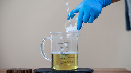 A person in blue gloves adds a solid substance into a measuring cup containing a yellow liquid,...