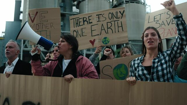 Nonconformist gen z activist woman shouting at a protest holding a megaphone to the group of people at a demonstration against pollution and factories with several banners of climate change messages