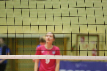 A volleyball net in an indoor court, with volleyball players blurred in the background