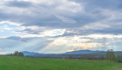 Bright blue sky over a calm emerald green meadow with rays of sunlight breaking through the clouds. The spring greenery contrasts with the bright sky.