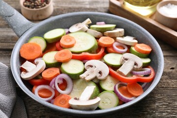 Frying pan with mix of fresh vegetables and mushrooms on wooden table, closeup