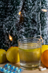 A festive arrangement of a glass of water with lemon, tablets, vitamins, and a Christmas tree in the background.