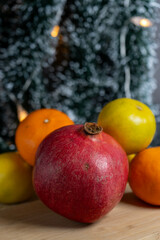 A composition of mandarins, lemons, and a pomegranate on a table with a Christmas tree and garlands in the background, highlighting festive abundance.
