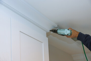 Carpenter is carefully installing crown molding in well lit room person uses power air gun to...