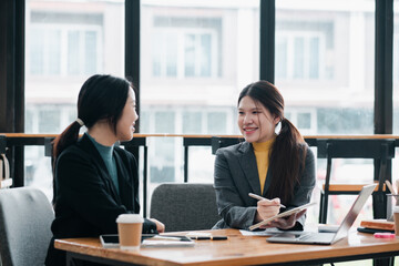 Two women in a modern office setting, discussing business strategies with laptops and notebooks.